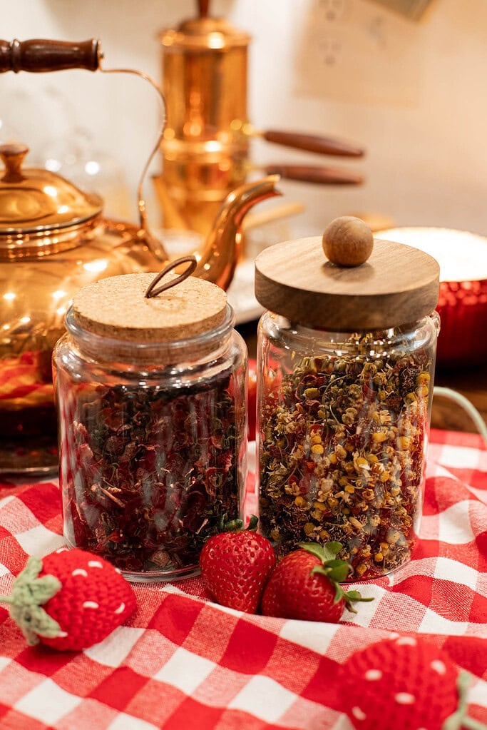 Glass jars of loose leaf strawberry tea ingredients on red gingham cloth with fresh strawberries and copper teapot in background