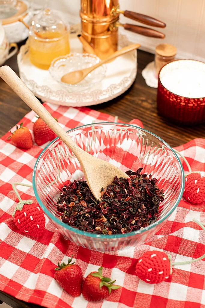 Loose leaf strawberry tea blend in glass bowl with wooden spoon on red gingham cloth with fresh strawberries
