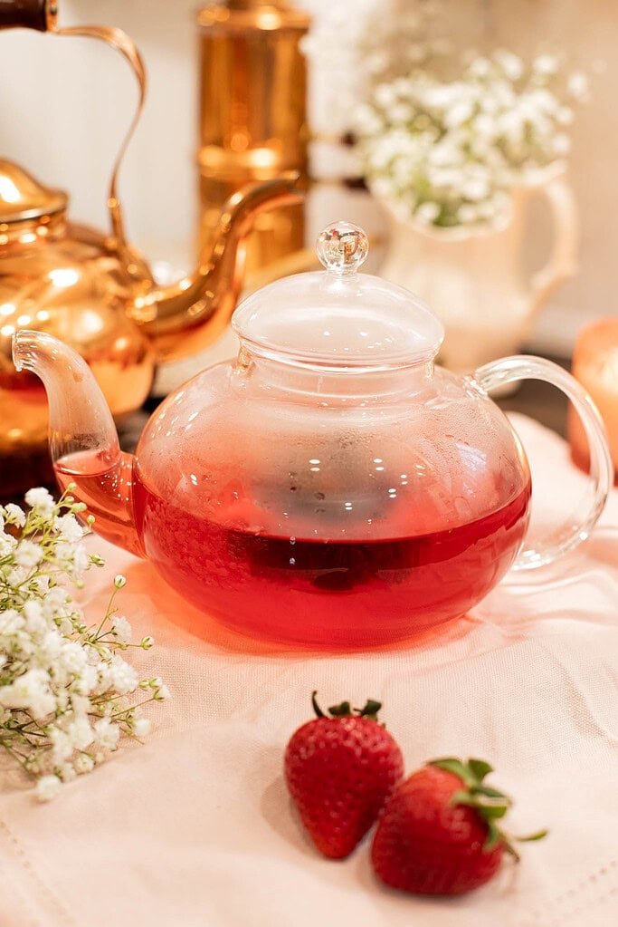 Glass teapot filled with freshly brewed strawberry tea on soft linen cloth with strawberries and copper kettle