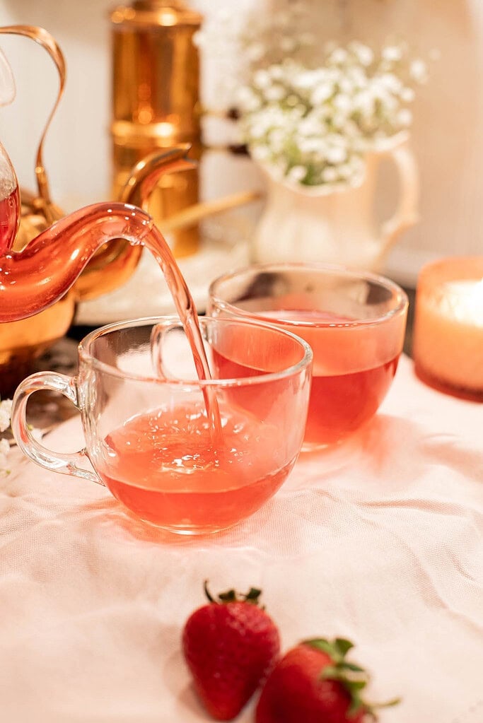 Pouring hot strawberry tea from glass teapot into clear tea cups with fresh strawberries nearby