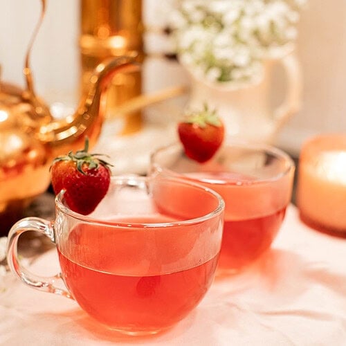 Two clear glass cups filled with strawberry tea garnished with fresh strawberries on cozy tea table