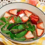 Top-down view of a summer simmer pot featuring strawberries, mint leaves, and vanilla bean pods in a white Dutch oven, styled with a yellow checkered cloth and a wooden bowl of fresh ingredients.