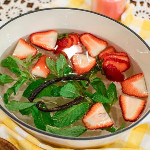 Top-down view of a summer simmer pot featuring strawberries, mint leaves, and vanilla bean pods in a white Dutch oven, styled with a yellow checkered cloth and a wooden bowl of fresh ingredients.