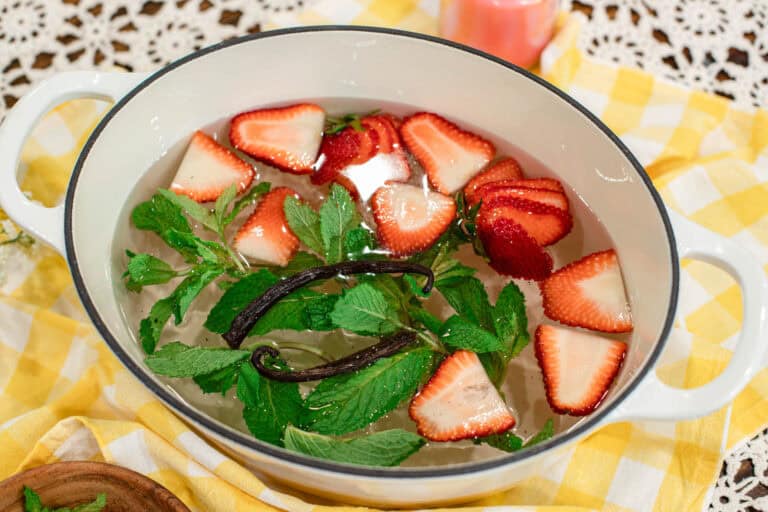 Top-down view of a summer simmer pot featuring strawberries, mint leaves, and vanilla bean pods in a white Dutch oven, styled with a yellow checkered cloth and a wooden bowl of fresh ingredients.