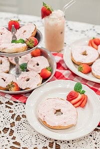Tiered tray of bagels topped with pink whipped strawberry cream cheese, garnished with fresh strawberries and mint, with an iced drink in the background.
