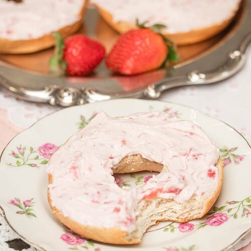 Strawberry cream cheese spread generously over a bagel on a floral plate with fresh strawberries in the background.