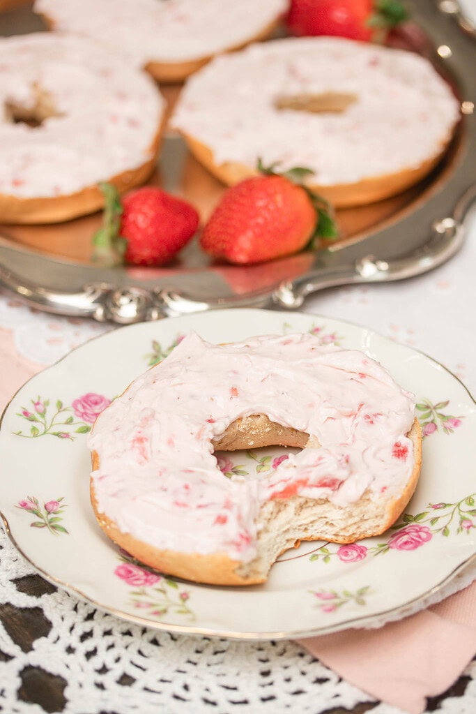 Strawberry cream cheese spread generously over a bagel on a floral plate with fresh strawberries in the background.