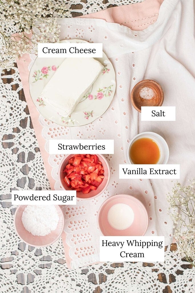 Overhead view of ingredients for whipped strawberry cream cheese including cream cheese, chopped strawberries, powdered sugar, vanilla extract, salt, and heavy whipping cream on a lace tablecloth.