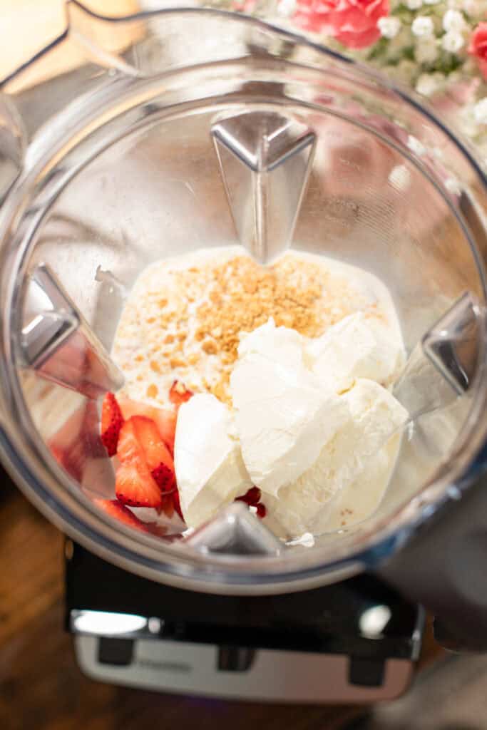 Top-down view of a blender filled with strawberries, cream cheese, crushed graham crackers, and milk, ready to be blended for strawberry cheesecake popsicles.