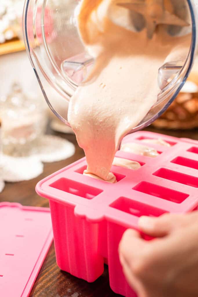 Creamy strawberry cheesecake mixture being poured from a blender into a pink silicone popsicle mold on a wooden counter.