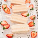 Four creamy strawberry cheesecake popsicles displayed on a white tray filled with ice, surrounded by halved strawberries on a lace tablecloth.