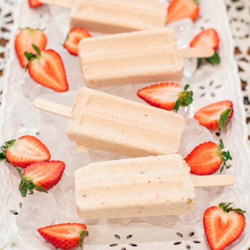 Four creamy strawberry cheesecake popsicles displayed on a white tray filled with ice, surrounded by halved strawberries on a lace tablecloth.