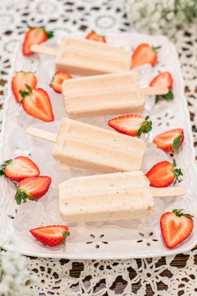 Four creamy strawberry cheesecake popsicles displayed on a white tray filled with ice, surrounded by halved strawberries on a lace tablecloth.