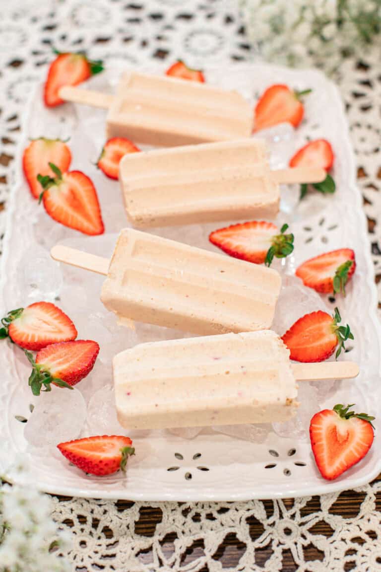 Four creamy strawberry cheesecake popsicles displayed on a white tray filled with ice, surrounded by halved strawberries on a lace tablecloth.