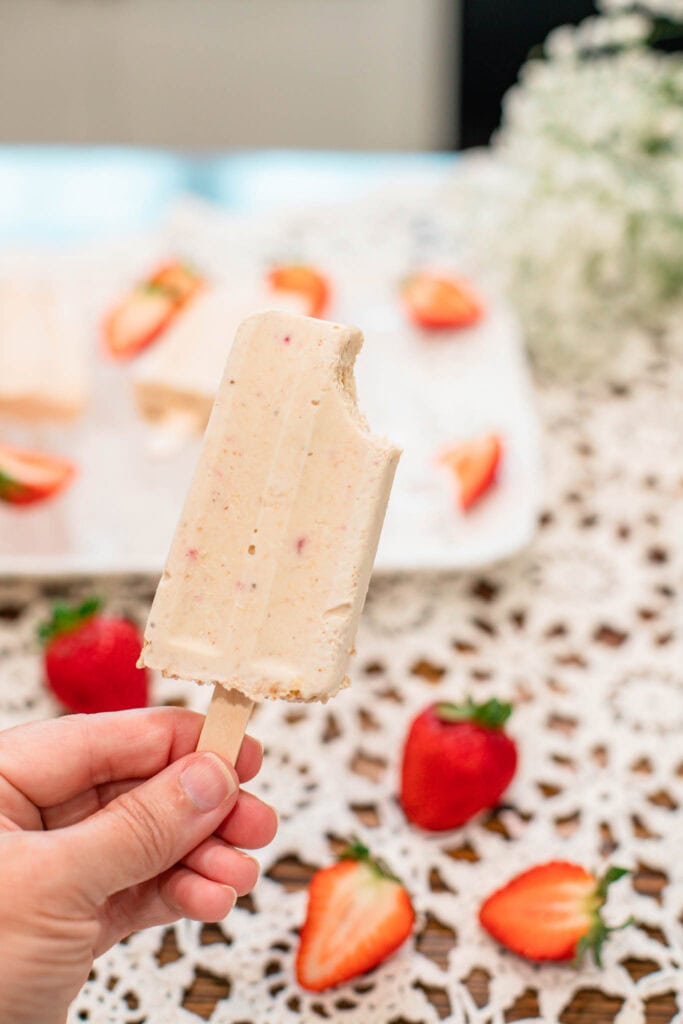 Hand holding one strawberry cheesecake popsicle with a bite taken out, with the tray of popsicles blurred in the background.