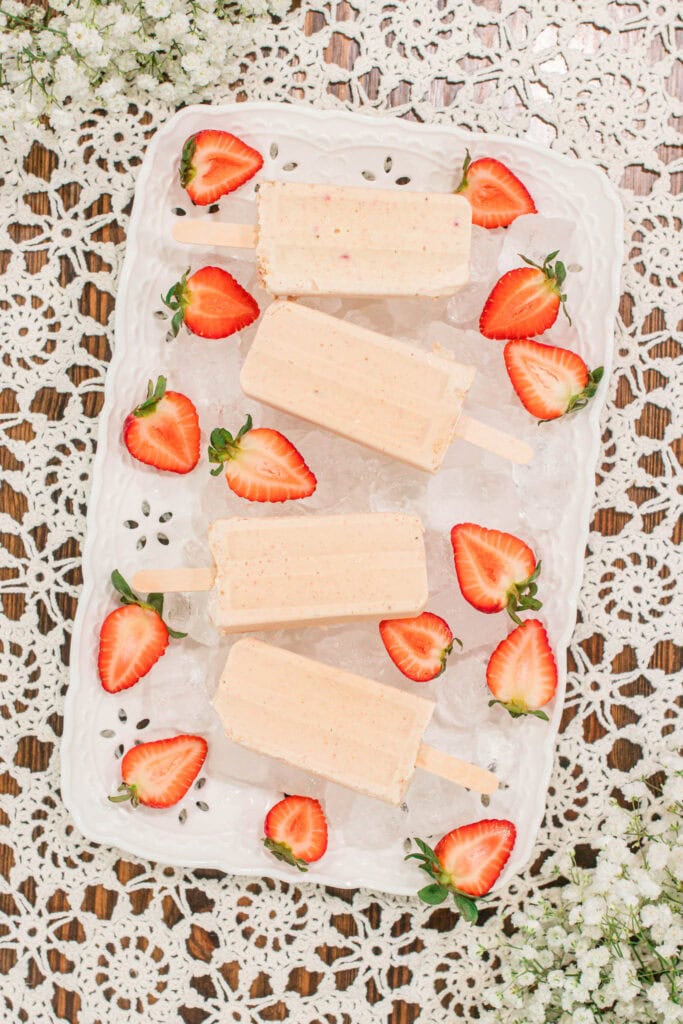 Overhead view of the popsicles and sliced strawberries arranged neatly on a white tray over a vintage lace table runner, with baby’s breath flowers framing the scene.