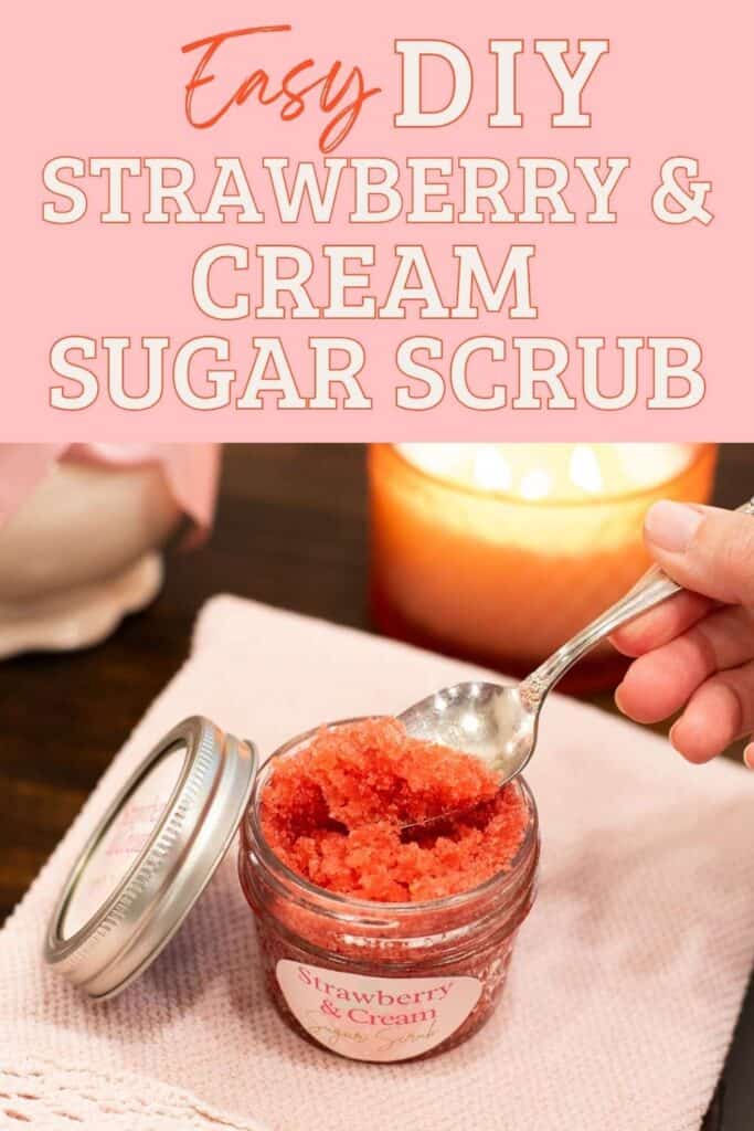 A small glass jar of homemade strawberry and cream sugar scrub sits on a soft pink cloth, with a spoon scooping out the bright red scrub. A glowing candle and a vase with pink ribbon are softly blurred in the background, creating a cozy, cottage-style atmosphere. Bold pink and white text at the top reads: “Easy DIY Strawberry & Cream Sugar Scrub.”