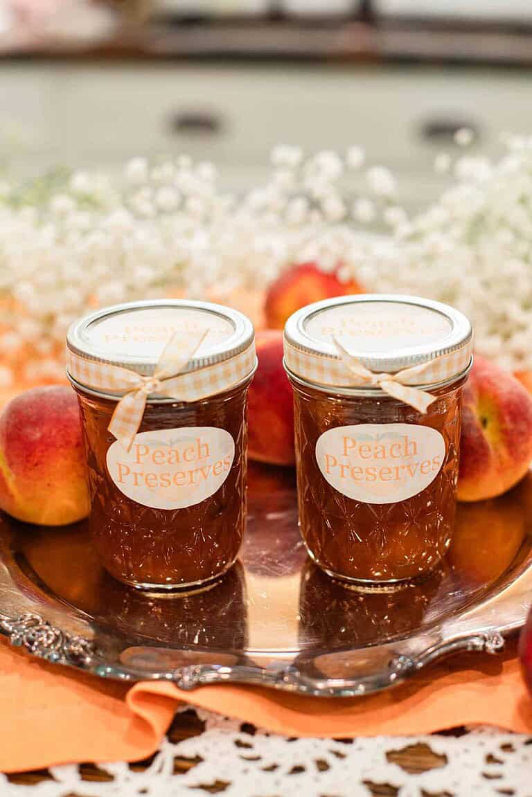 Two mason jars labeled “Peach Preserves” with gingham ribbons, surrounded by fresh peaches and baby’s breath flowers on a silver tray.