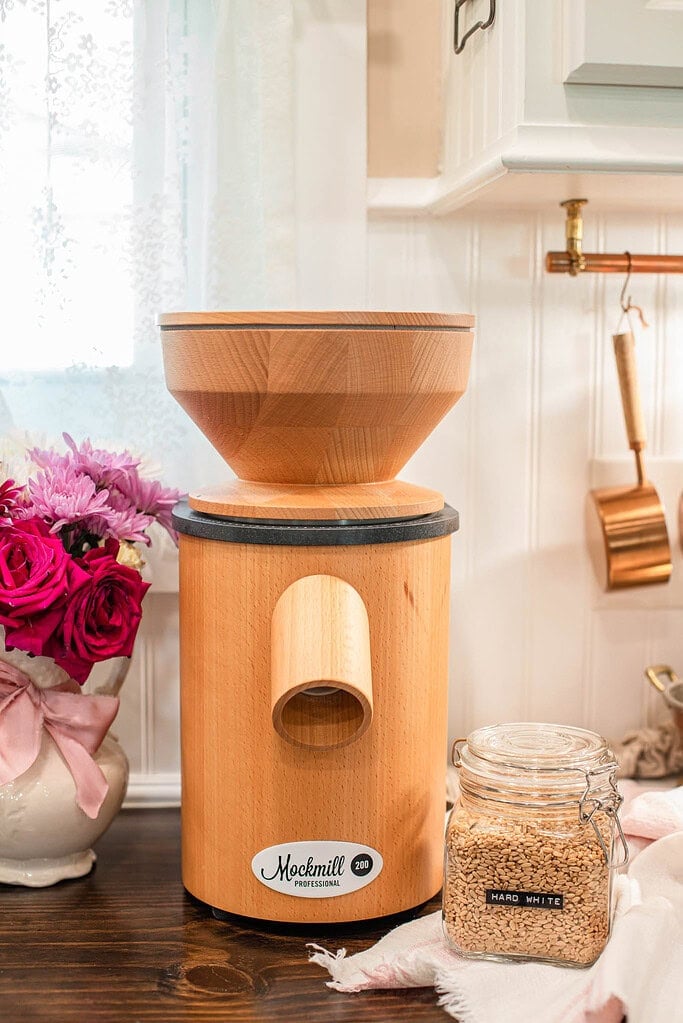 Wooden grain mill on a kitchen counter next to a glass jar of hard white wheat berries and a vase of pink and red flowers in a bright cottage-style kitchen