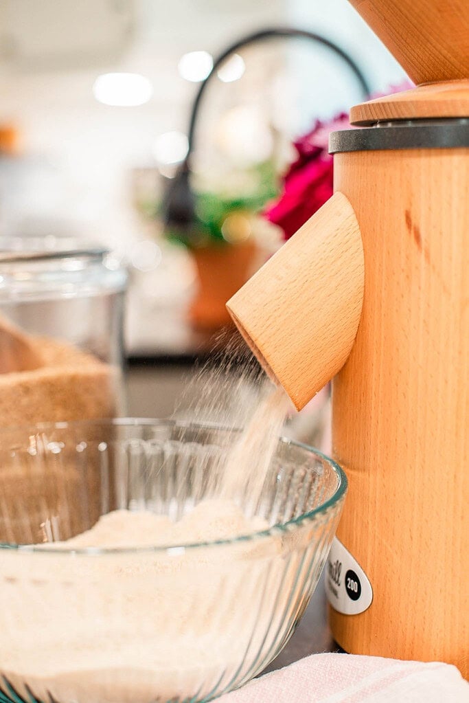 Freshly milled flour pouring from a wooden grain mill spout into a clear glass mixing bowl