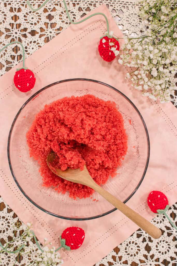 A glass bowl filled with vibrant strawberry and cream sugar scrub, placed on a soft pink cloth with a wooden spoon resting inside. Crocheted strawberries and delicate white baby’s breath flowers add a whimsical cottage touch.