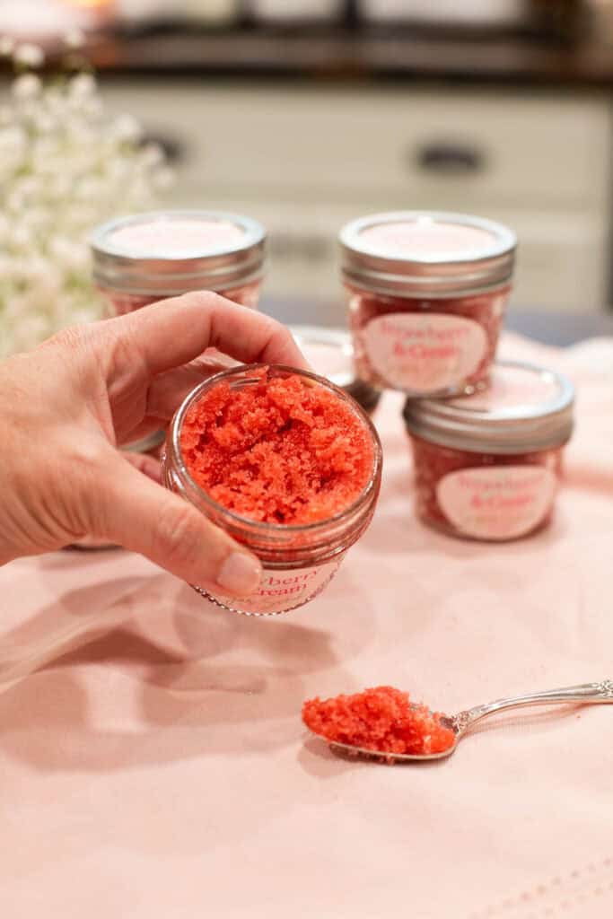A hand holding a small glass jar of strawberry and cream sugar scrub, with a spoonful of the scrub in the foreground. In the background, additional labeled jars sit on a pink cloth, suggesting a homemade gift or self-care project.