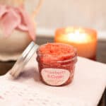 A cozy scene featuring a jar of strawberry and cream sugar scrub on a folded pink knit cloth. A soft candle glows in the background next to a pitcher tied with a pink ribbon and filled with white flowers, evoking calm and comfort.