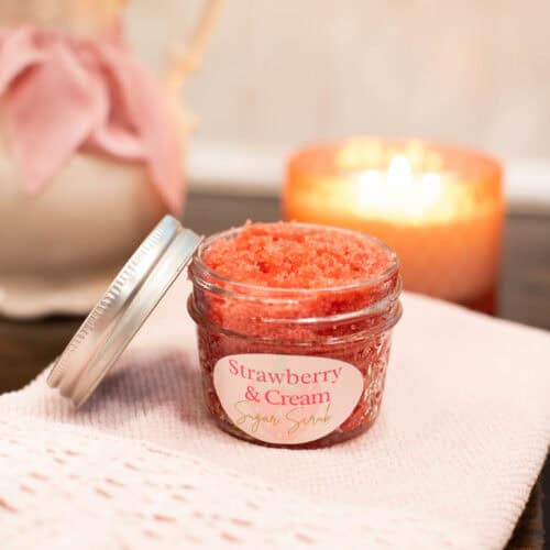 A cozy scene featuring a jar of strawberry and cream sugar scrub on a folded pink knit cloth. A soft candle glows in the background next to a pitcher tied with a pink ribbon and filled with white flowers, evoking calm and comfort.