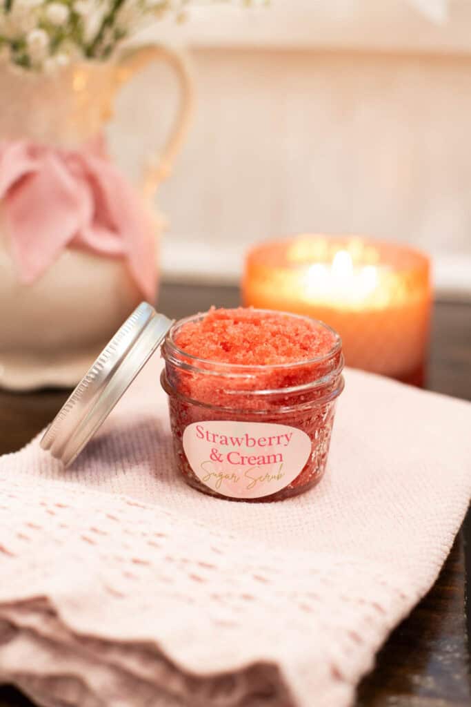 A cozy scene featuring a jar of strawberry and cream sugar scrub on a folded pink knit cloth. A soft candle glows in the background next to a pitcher tied with a pink ribbon and filled with white flowers, evoking calm and comfort.