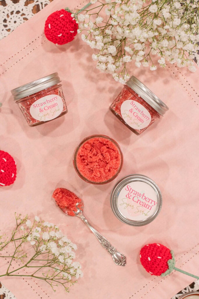 A flat lay of several jars of strawberry and cream sugar scrub arranged on a pink linen with baby’s breath, crocheted strawberries, and a vintage-style spoon holding a scoop of the scrub. A charming display of cottage-style homemade skincare.