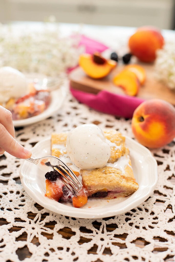 Fork cutting into sourdough peach blackberry galette with vanilla ice cream and juicy fruit filling