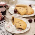 A fork lifting a bite of flaky cherry-filled hand pie from a floral plate, with more pies and fresh cherries in the background.