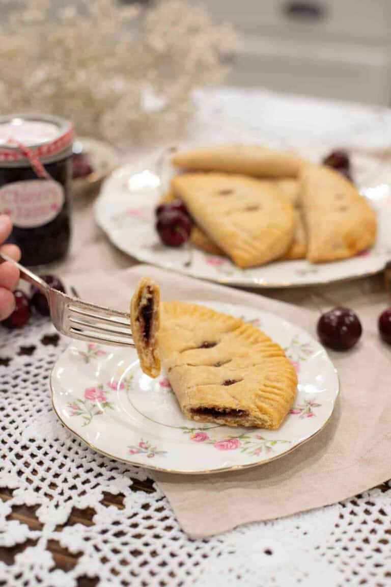 A fork lifting a bite of flaky cherry-filled hand pie from a floral plate, with more pies and fresh cherries in the background.