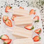 Strawberry cheesecake popsicles on a white decorative tray filled with ice and surrounded by fresh sliced strawberries on a lace tablecloth.