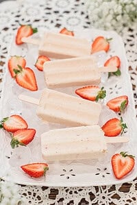 Strawberry cheesecake popsicles on a white decorative tray filled with ice and surrounded by fresh sliced strawberries on a lace tablecloth.