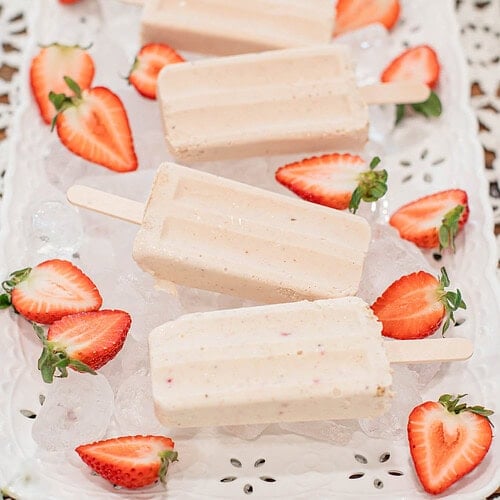 Strawberry cheesecake popsicles on a white decorative tray filled with ice and surrounded by fresh sliced strawberries on a lace tablecloth.