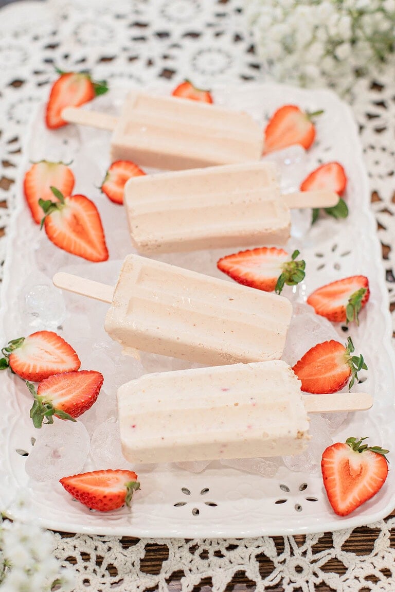 Strawberry cheesecake popsicles on a white decorative tray filled with ice and surrounded by fresh sliced strawberries on a lace tablecloth.