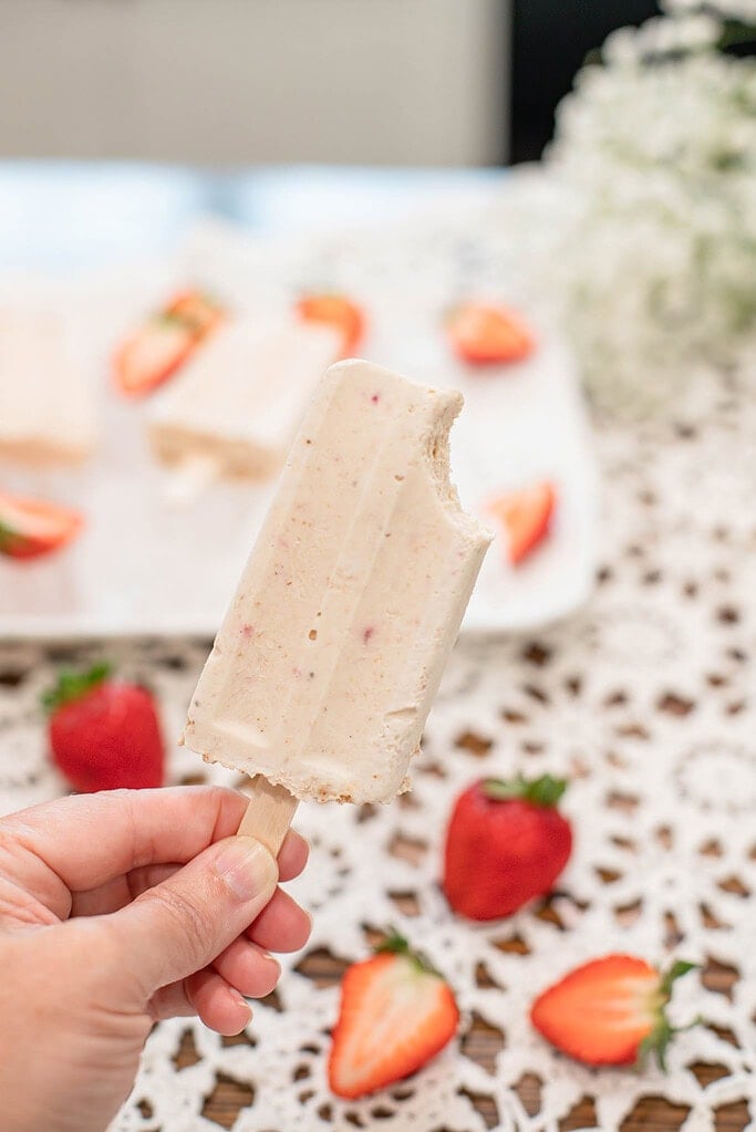 Hand holding a strawberry cheesecake popsicle with a bite taken out, fresh strawberries blurred in the background.