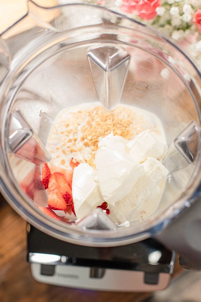 Blender filled with strawberries, cream cheese, cream, and crushed graham crackers before blending.