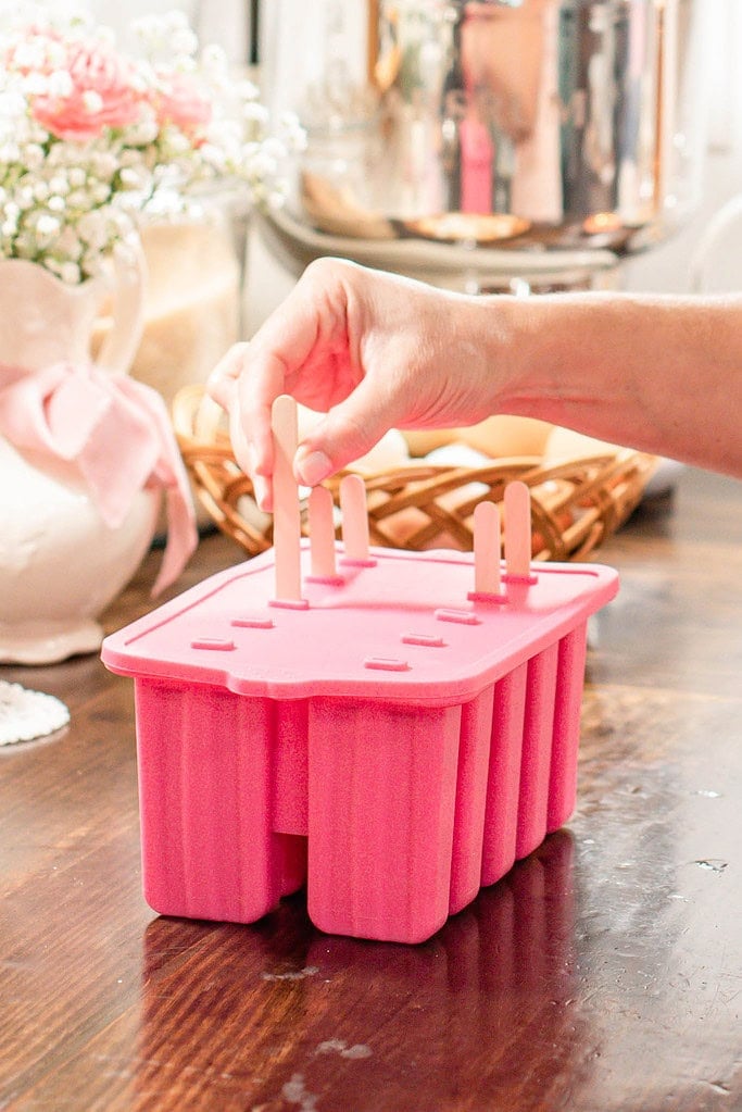 Hand inserting wooden popsicle sticks into a pink silicone popsicle mold filled with strawberry cheesecake mixture.