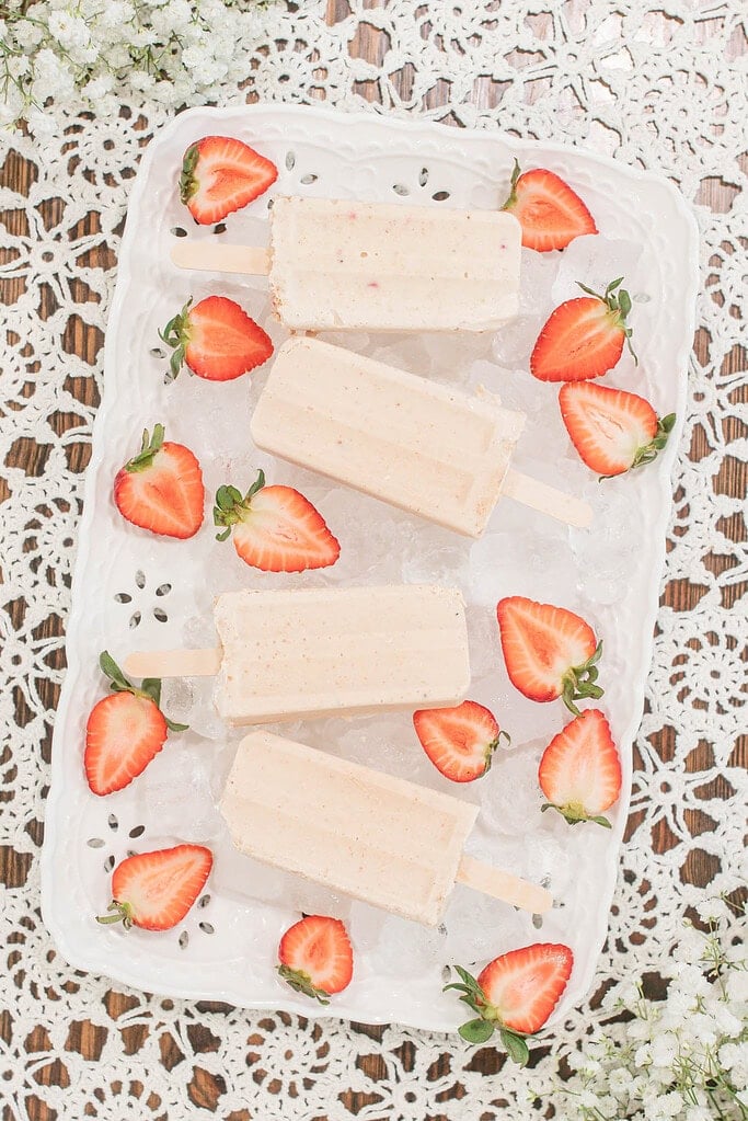 Overhead view of strawberry cheesecake popsicles on ice with fresh strawberries arranged around them on a white tray.