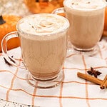 Two clear glass mugs of pumpkin spice latte with whipped cream topping, placed on a festive fall table with pumpkins, cinnamon sticks, and a copper tea kettle in the background.