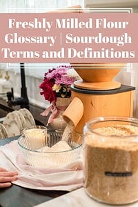 Freshly milled flour being ground into a glass bowl from a countertop grain mill in a cozy kitchen setting, with wheat berries in a jar and flowers nearby, and text overlay reading “Freshly Milled Flour Glossary | Sourdough Terms and Definitions.”