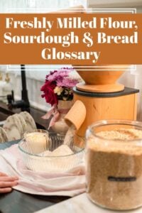 Freshly milled flour being poured from a wooden grain mill into a glass mixing bowl on a cottage kitchen table, with a jar of wheat berries and a vase of flowers nearby. Text overlay reads ‘Freshly Milled Flour, Sourdough & Bread Glossary’