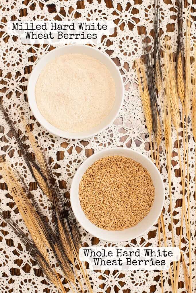 Side-by-side comparison of whole hard white wheat berries and freshly milled hard white wheat flour in white bowls, styled on a lace tablecloth with wheat stalks.