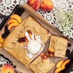 Freshly baked sourdough blackberry peach galette on a parchment-lined cast iron pan, topped with a scoop of vanilla ice cream, surrounded by fresh peach slices, blackberries, and sprigs of white flowers on a lace tablecloth.