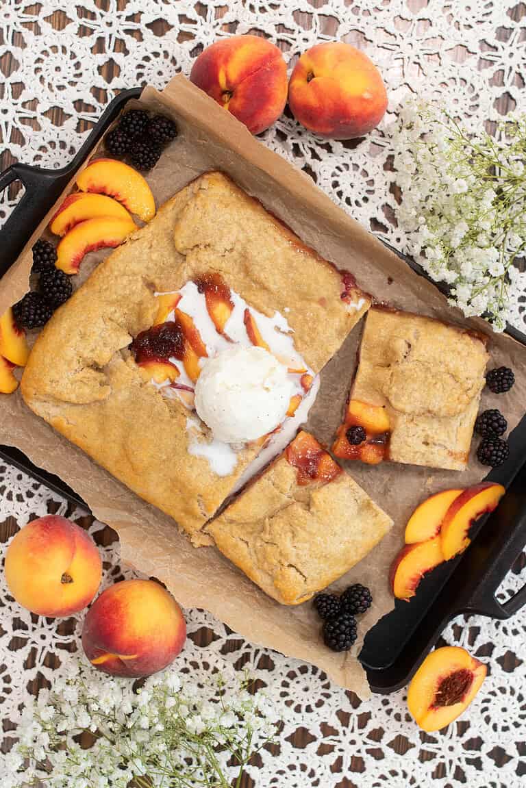 Freshly baked sourdough blackberry peach galette on a parchment-lined cast iron pan, topped with a scoop of vanilla ice cream, surrounded by fresh peach slices, blackberries, and sprigs of white flowers on a lace tablecloth.