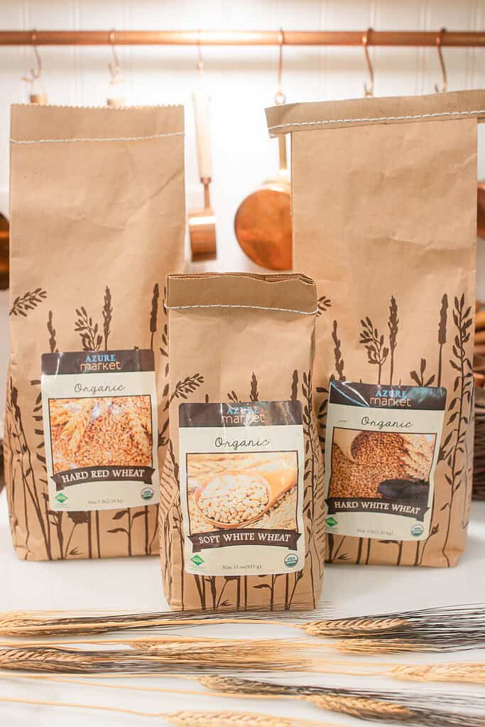 Three brown paper bags of organic wheat berries from Azure Market, labeled Hard Red Wheat, Soft White Wheat, and Hard White Wheat, displayed on a kitchen counter with dried wheat stalks in front and copper kitchen utensils hanging in the background.