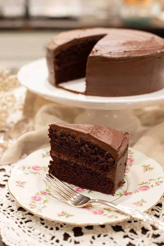 A slice of rich chocolate layer cake with frosting on a floral china plate, with the rest of the cake on a stand in the background.