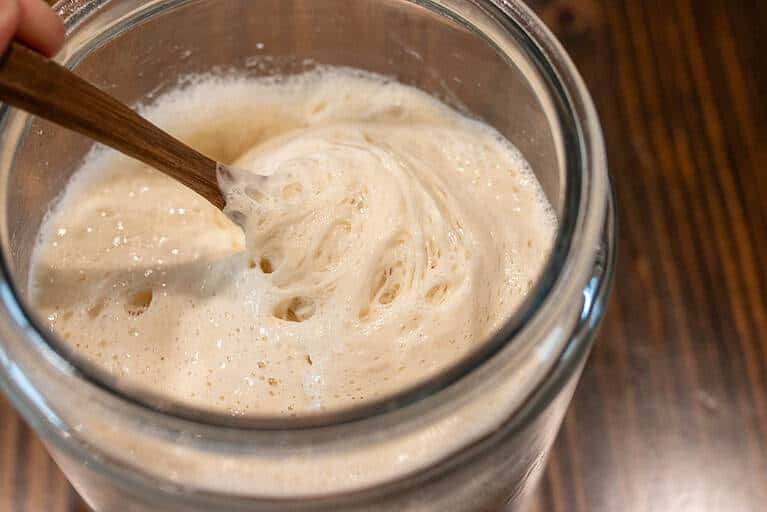 A glass jar of active sourdough starter with bubbles and a spoon inside.